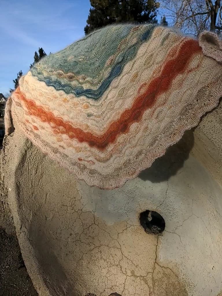 A close-up photograph of a hand-knitted shawl draped over a textured, light-colored surface outdoors. The shawl features horizontal stripes in shades of cream, rust orange, and blue-green, with a scalloped lace edge.