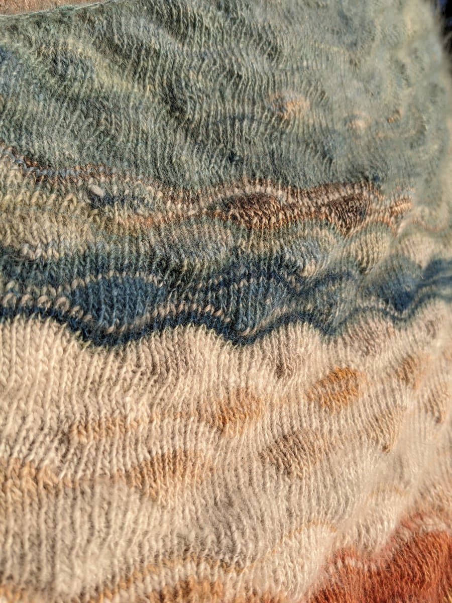 A close-up photograph of a hand-knitted textile featuring horizontal stripes in earthy tones of blue, green, cream, and rust. The image highlights the texture of the yarn and the distinct stitch patterns used in each color section.