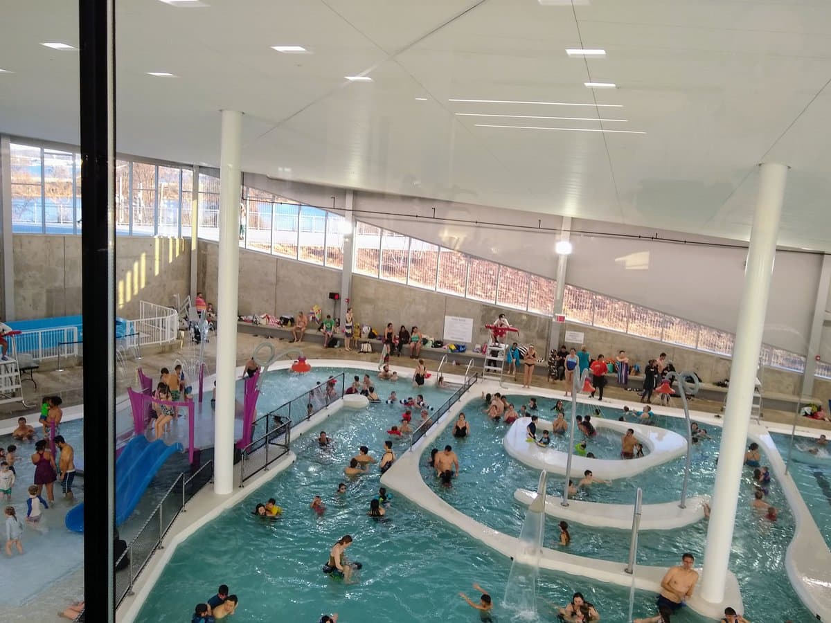 A wide-angle photograph of a busy indoor aquatic center, showing a large pool with a winding lazy river, a children's play area with a blue slide, and numerous people swimming and relaxing on the deck. The facility features a high, white paneled ceiling, large windows along the upper walls, and white support columns.