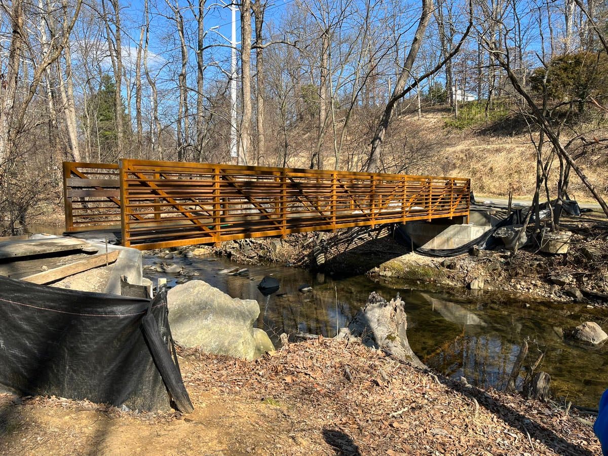 A newly constructed wooden footbridge with horizontal railings spans a small, rocky stream in a wooded area. The surrounding landscape features bare trees and dry leaves, indicating a late autumn or early spring setting.