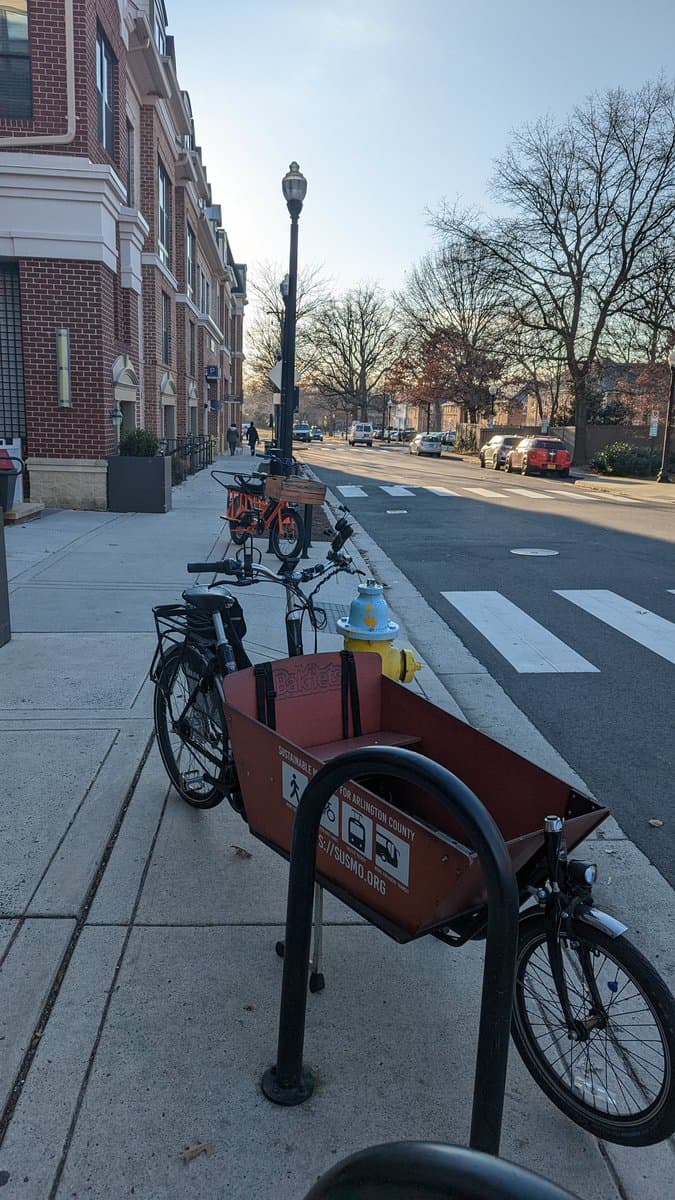 A red cargo bike is parked on a city sidewalk next to a bike rack, with a blue and yellow fire hydrant and a brick building in the background. Another orange cargo bike is visible further down the sidewalk.