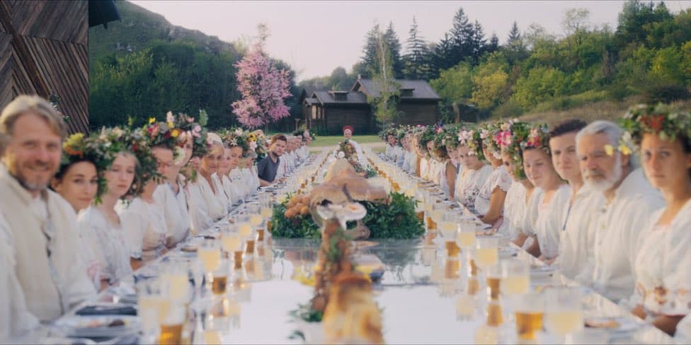 A wide shot of a long outdoor banquet table set in a forest clearing, with numerous people wearing white clothing and floral wreaths seated on both sides. The table is decorated with a large centerpiece featuring a whole roasted animal and greenery, with glasses of beer or juice in front of each guest.