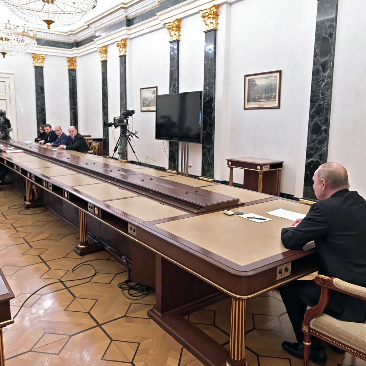 A photograph of a formal meeting room featuring a very long, dark wooden conference table. A man in a dark suit sits alone at the near end of the table, while a group of four men sits at the far end, with a professional video camera positioned between them.