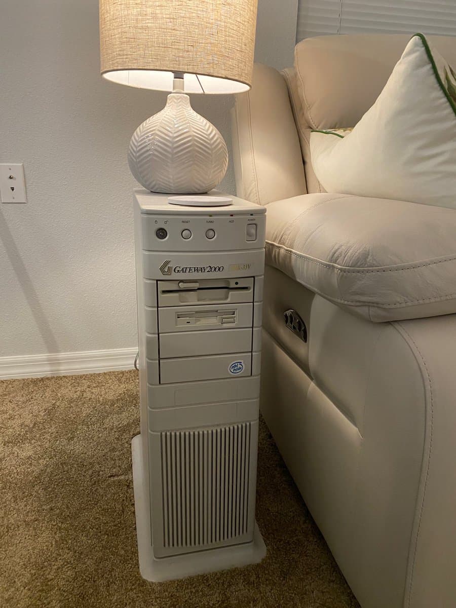 A photograph of a white Gateway 2000 desktop computer tower standing on a carpeted floor next to a white leather sofa, being used as an end table with a lamp placed on top of it.