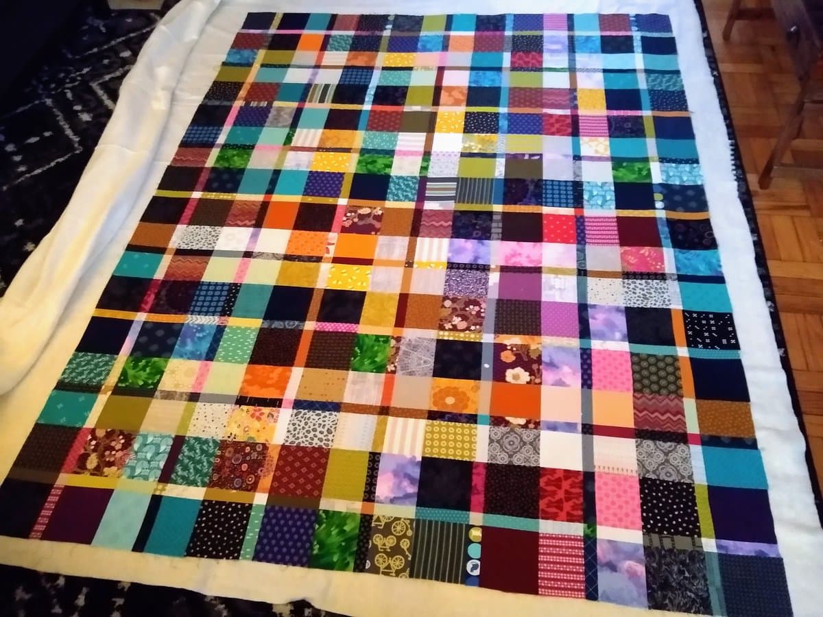A colorful patchwork quilt made of many small, multi-colored fabric squares is spread out on a white surface, likely a bed, in a room with a wooden floor.