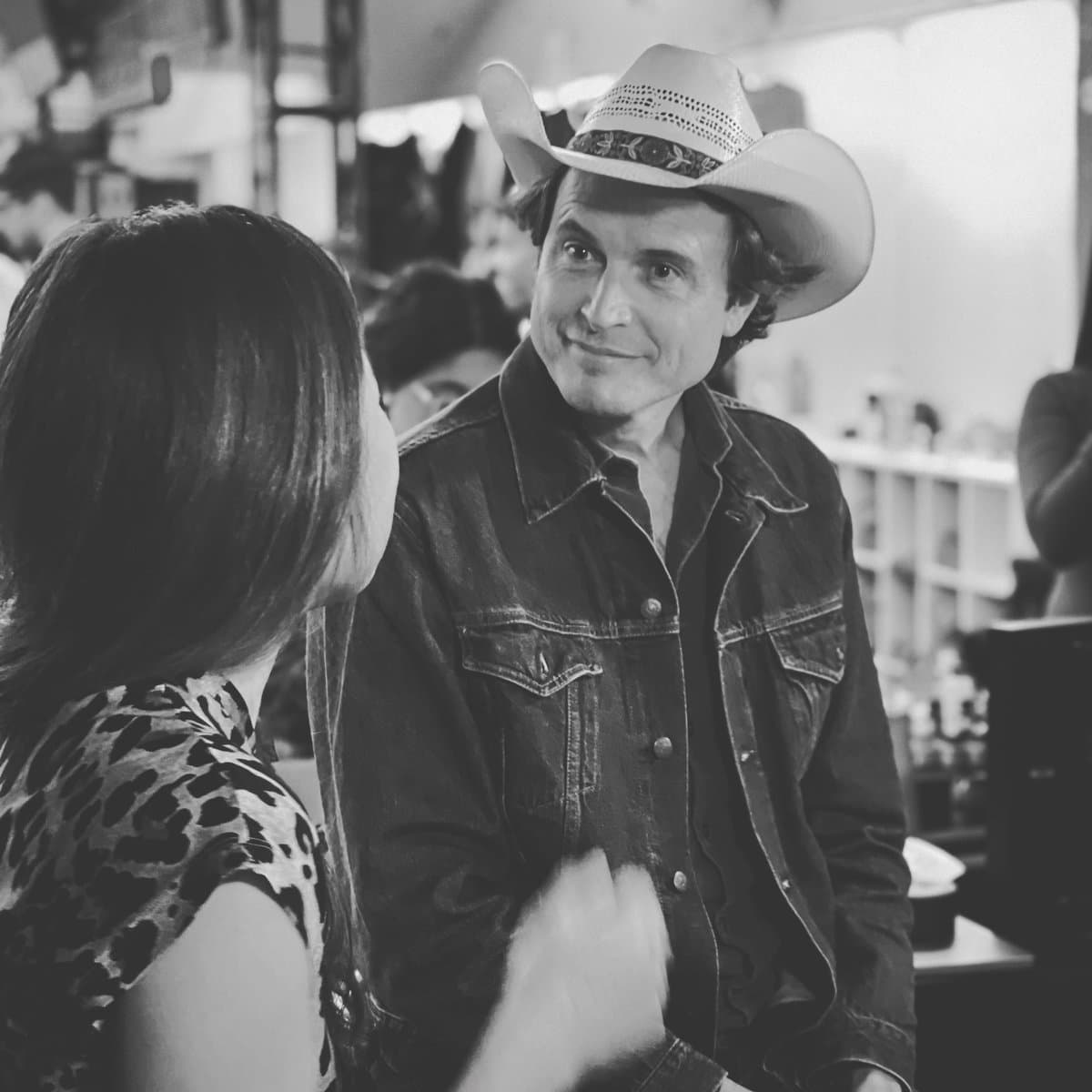 A black-and-white photograph showing a man wearing a cowboy hat and denim jacket smiling while looking at a woman with her back to the camera.