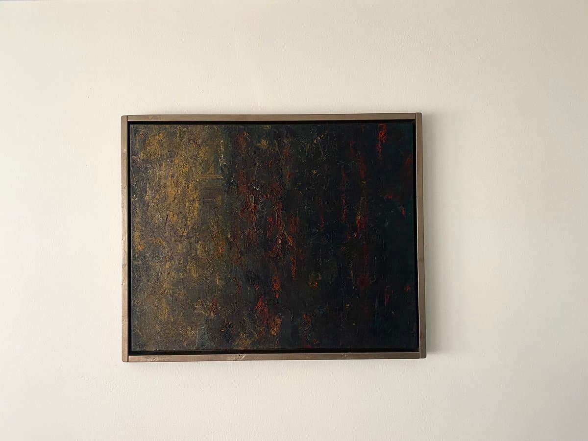 A framed abstract painting hangs on a plain white wall. The artwork features a dark, textured surface with vertical streaks of black, brown, and red, enclosed in a simple metallic frame.