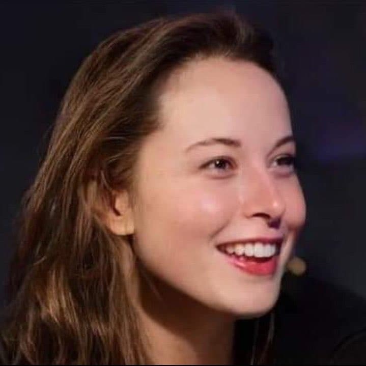 A close-up photograph of a smiling woman with long brown hair, shown in profile against a dark background. She is wearing a small lavalier microphone clipped to her clothing. The image is a standard portrait photo, not a text-bearing graphic.
