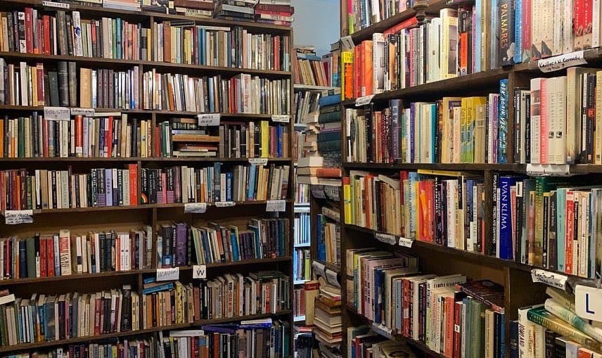 A photograph of a densely packed bookshelf in a library or bookstore, filled with numerous books of various sizes and colors. The shelves are organized with small white labels indicating sections or categories, and the books are arranged tightly together.