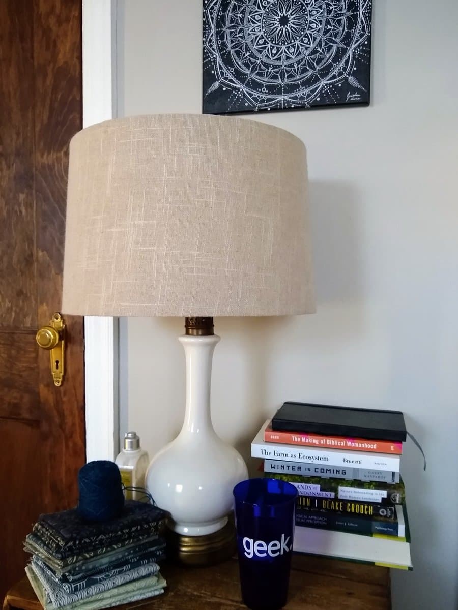 A photo of a cluttered nightstand featuring a white ceramic lamp, a stack of folded fabric, a blue 'geek' tumbler, and a pile of books. A black and white mandala print hangs on the wall above the lamp.