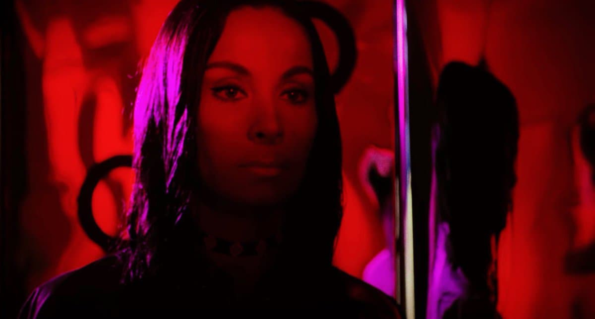 A still from the film 'The Other Side of the Wind' (2018) featuring a woman with dark hair and a choker necklace, illuminated by intense red and purple lighting. She is looking directly at the camera with a serious expression, while a mirror to her right reflects her profile.