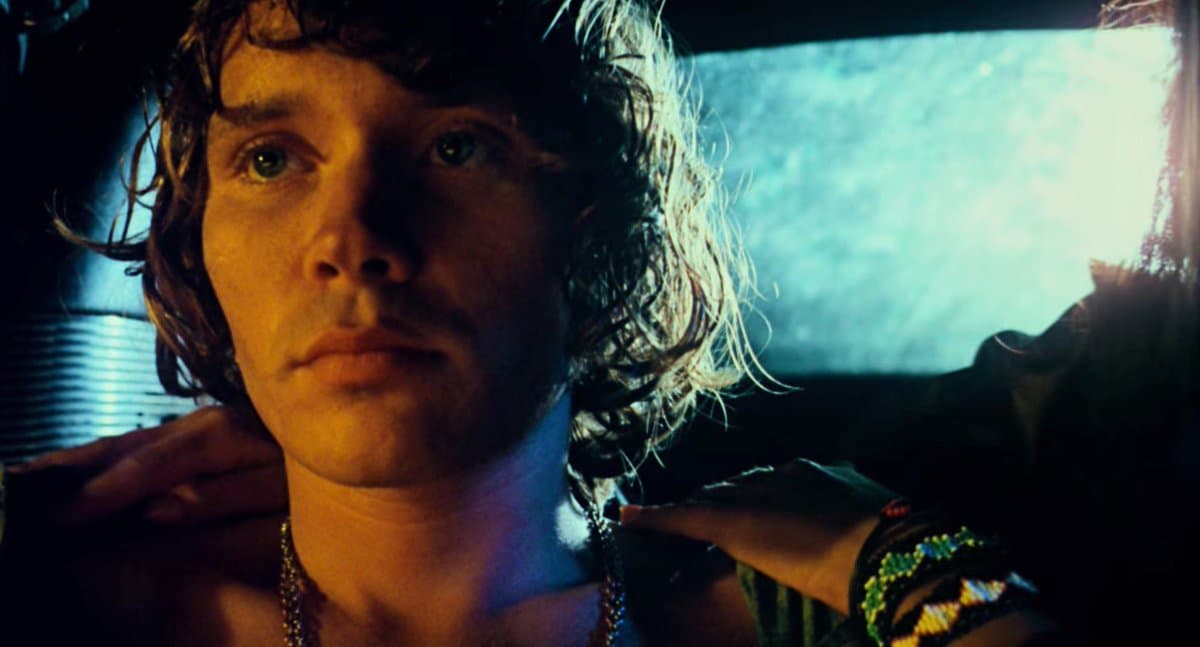 A close-up still from the film 'The Other Side of the Wind' (2018) featuring a man with curly hair looking directly at the camera, illuminated by warm and cool lighting. Another person's hands are visible on either side of his shoulders, with one hand wearing a beaded bracelet.