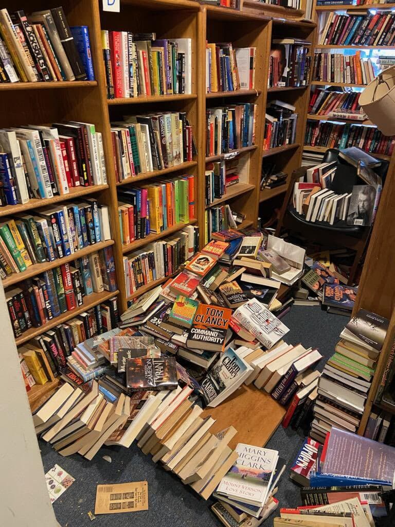 A photograph of a bookshelf in a library or bookstore that has been knocked over, spilling a large pile of books onto the floor. The scene shows wooden shelves packed with books, with a significant portion of the collection now scattered in a heap on the carpeted floor in front of the damaged shelving unit.