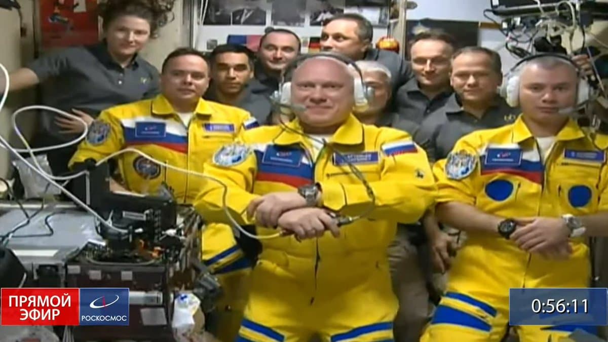 A group of astronauts in bright yellow flight suits and mission patches pose for a photo inside a spacecraft, with several crew members in dark shirts standing behind them. The image is a screenshot from a broadcast, indicated by the on-screen graphics.