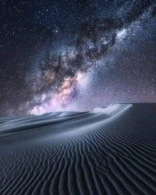 A long-exposure photograph of the Milky Way galaxy arching across a star-filled night sky above a rippled sand dune in a desert landscape. The foreground shows wind-carved patterns in the sand, illuminated by moonlight or ambient light, while the sky displays dense star fields and the bright, colorful core of the galaxy.