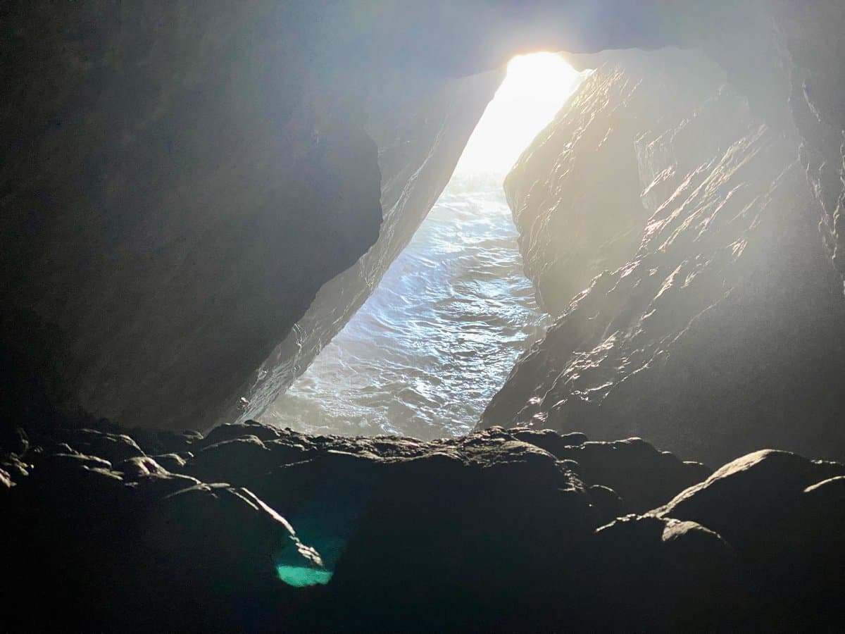 A view from inside a dark sea cave looking out toward the ocean, with bright sunlight streaming through the opening between the rock walls.