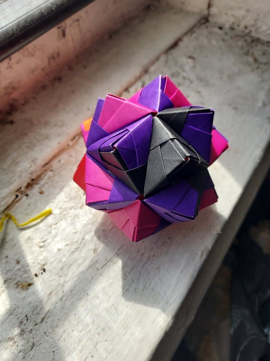A colorful, multi-faceted origami sphere made of folded paper in shades of purple, pink, and black, resting on a weathered white windowsill.