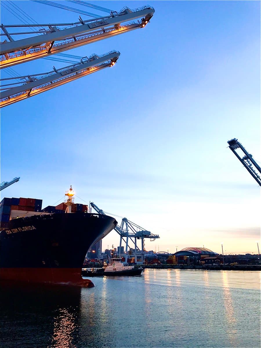 A photograph of a large container ship named 'CMA CGM FLORIDA' docked at a port terminal during twilight, with a tugboat nearby and large loading cranes overhead.