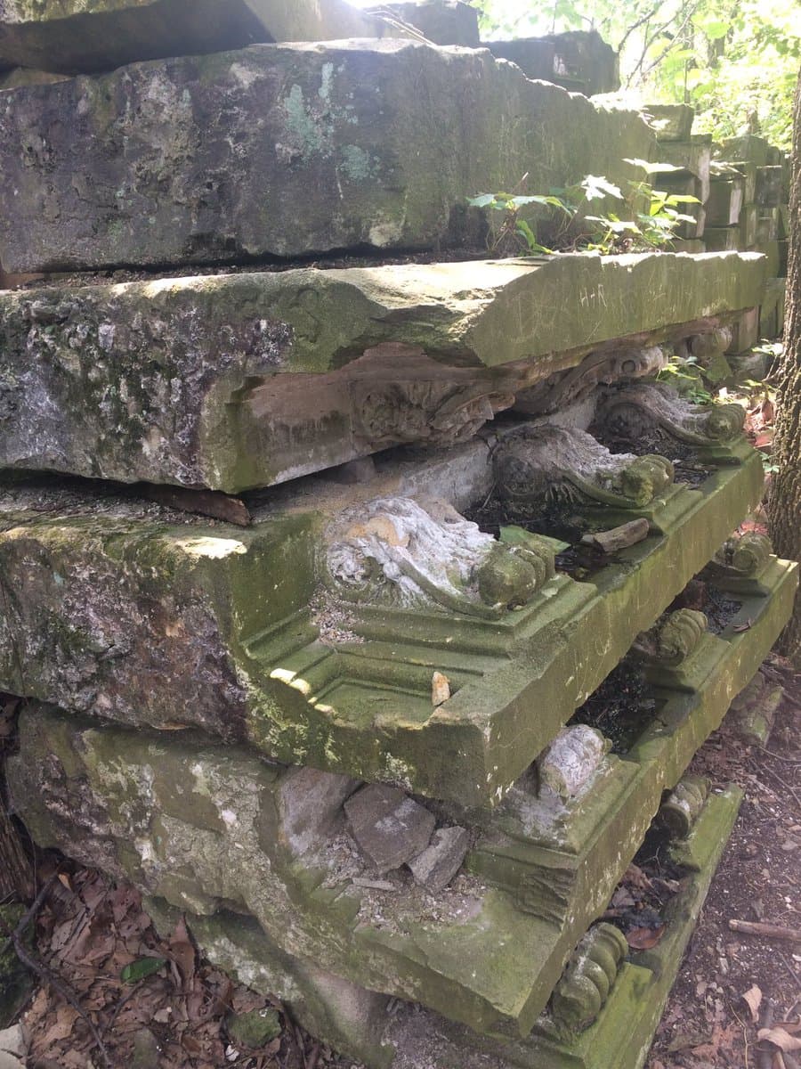 A close-up photograph of stacked, moss-covered stone architectural fragments, including carved capitals and bases, resting on a forest floor.