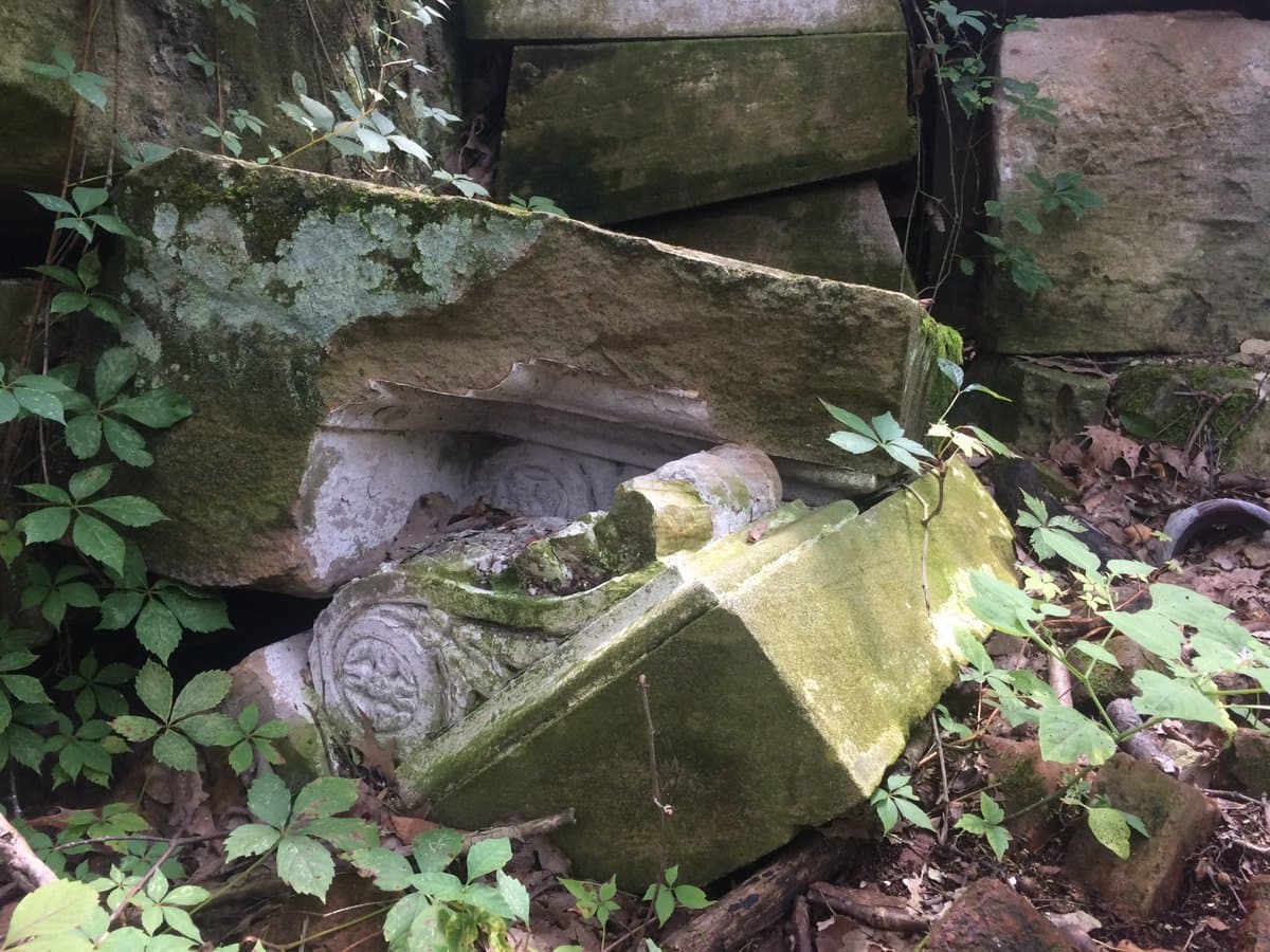 A close-up photograph of large, moss-covered stone blocks and a carved column fragment lying on the ground among green foliage and fallen leaves. The weathered pieces appear to be architectural remnants, with one block showing a carved spiral or scroll design.