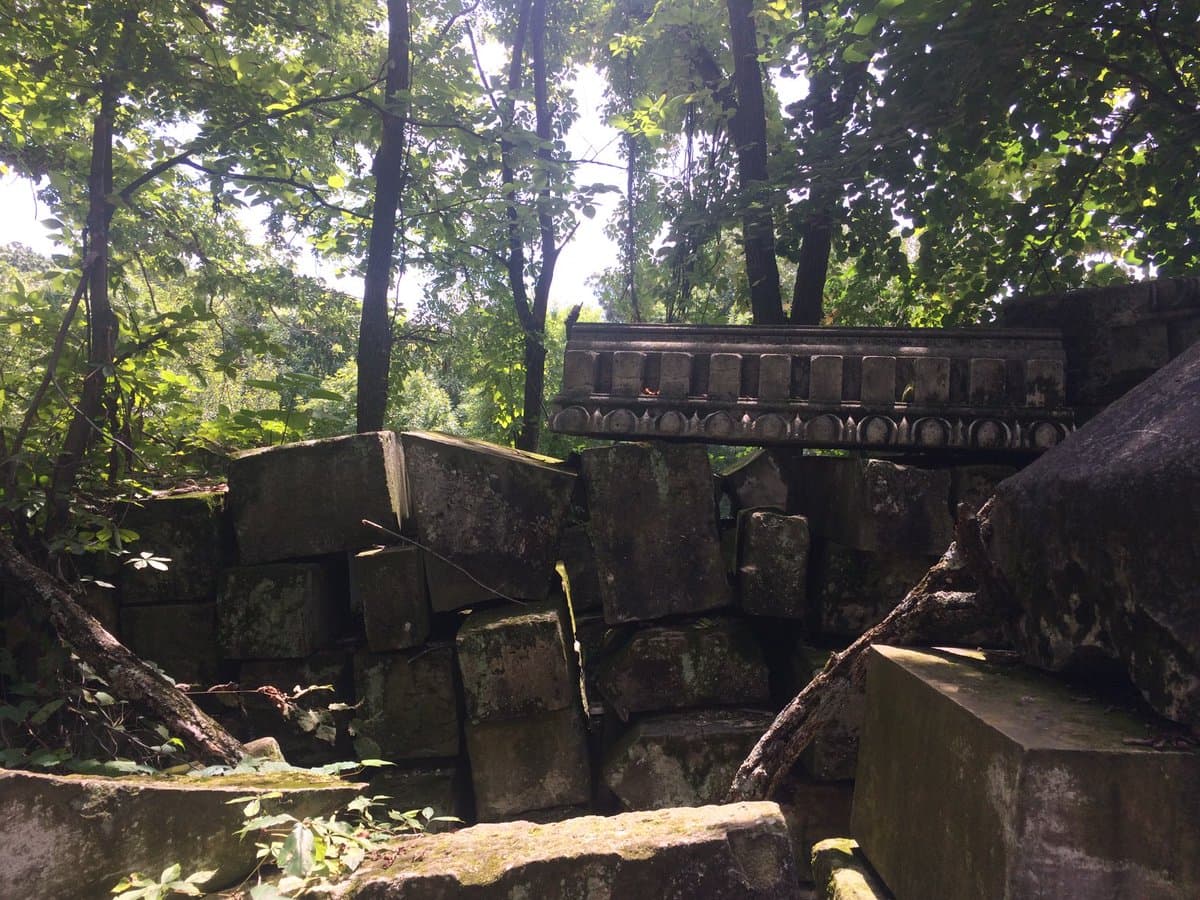 A photograph showing the weathered stone remains of a column base and entablature, including a row of carved blocks, situated in a forest clearing.