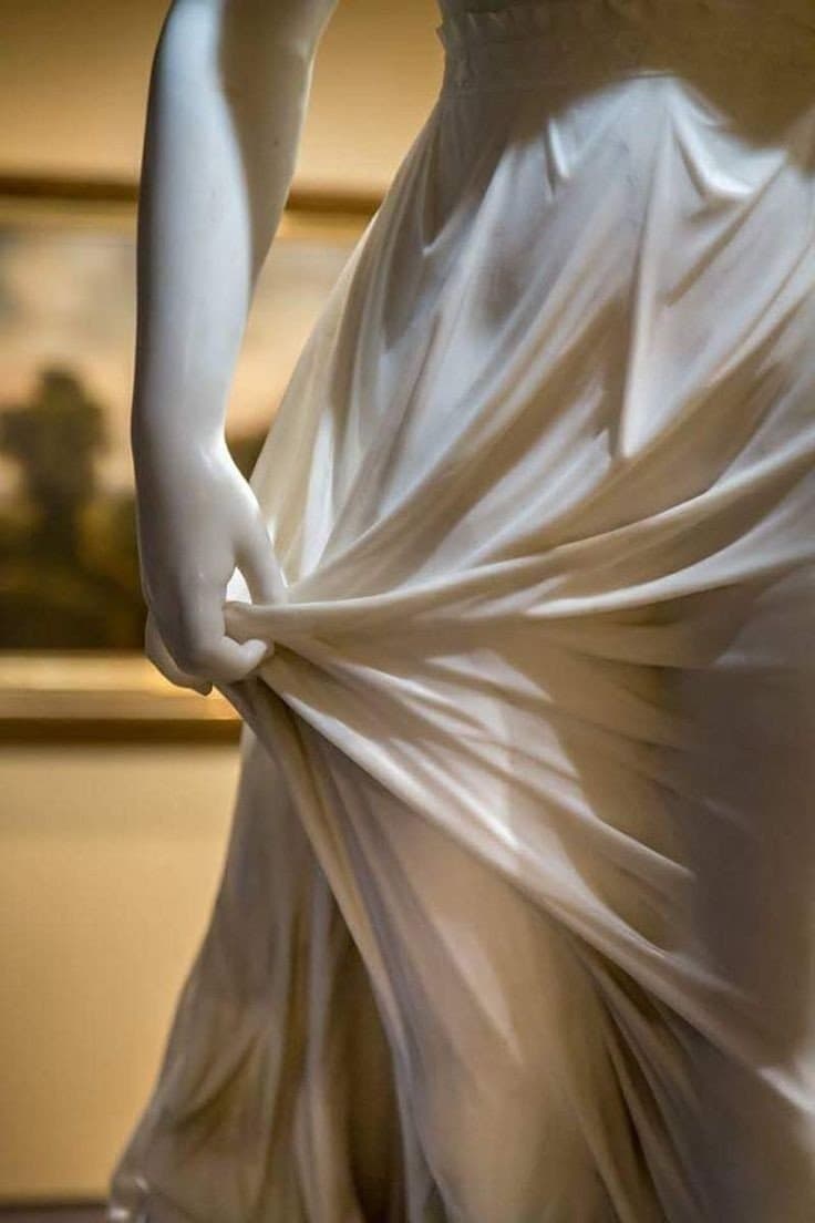 A close-up photograph of a white marble statue showing a figure's arm and hand pulling at the fabric of a draped garment. The background is a blurred interior space, likely a museum.