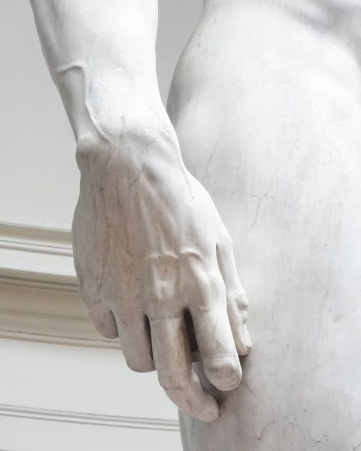 A close-up photograph of a white marble statue's hand and forearm, showing detailed veins and fingers resting against the figure's thigh. The image is a photo of a sculpture, not a text-bearing image.