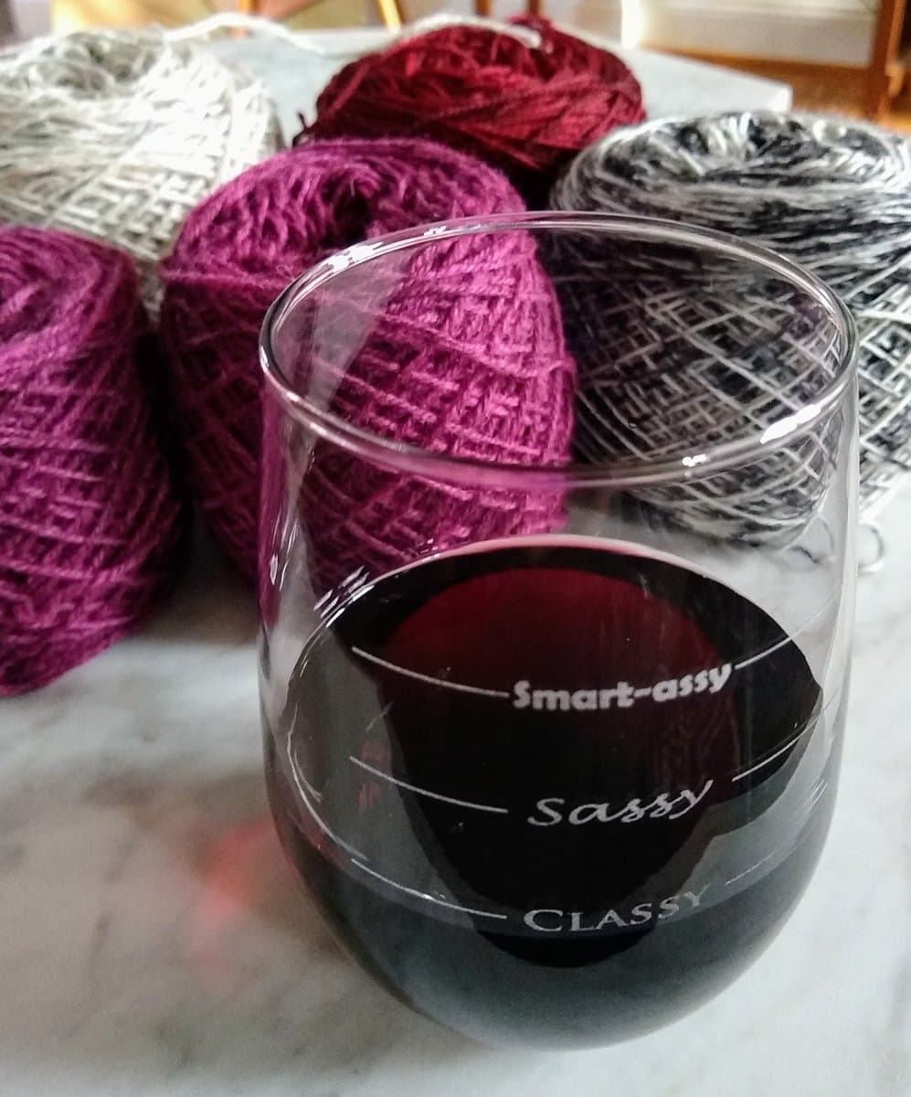 A close-up photograph of a glass of red wine sitting on a marble surface in front of several balls of colorful yarn. The glass features a humorous scale printed on the side.