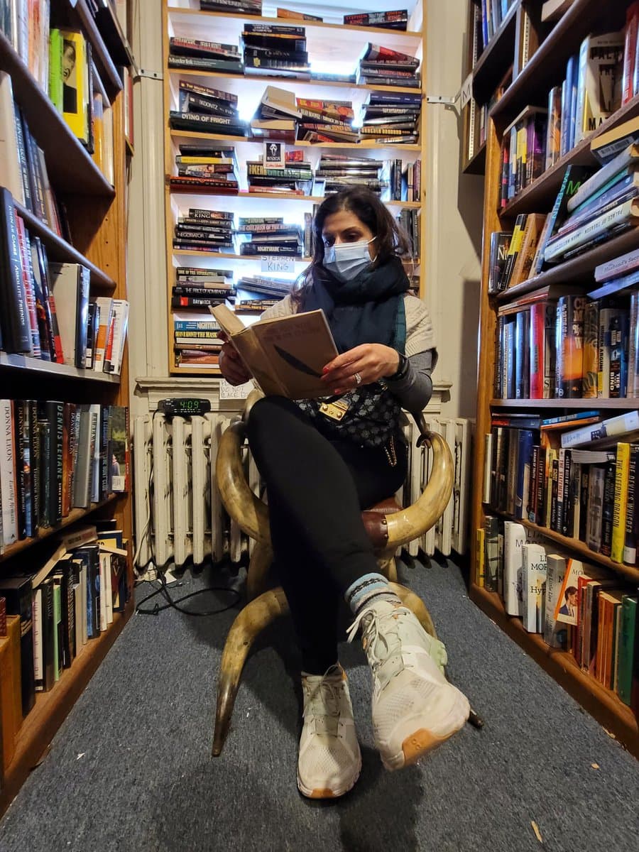 A woman wearing a face mask sits on a chair made of large animal horns, reading a book in a narrow aisle between tall, packed bookshelves in a bookstore. She is dressed in a sweater, scarf, and sneakers, with a digital clock visible on a radiator behind her.