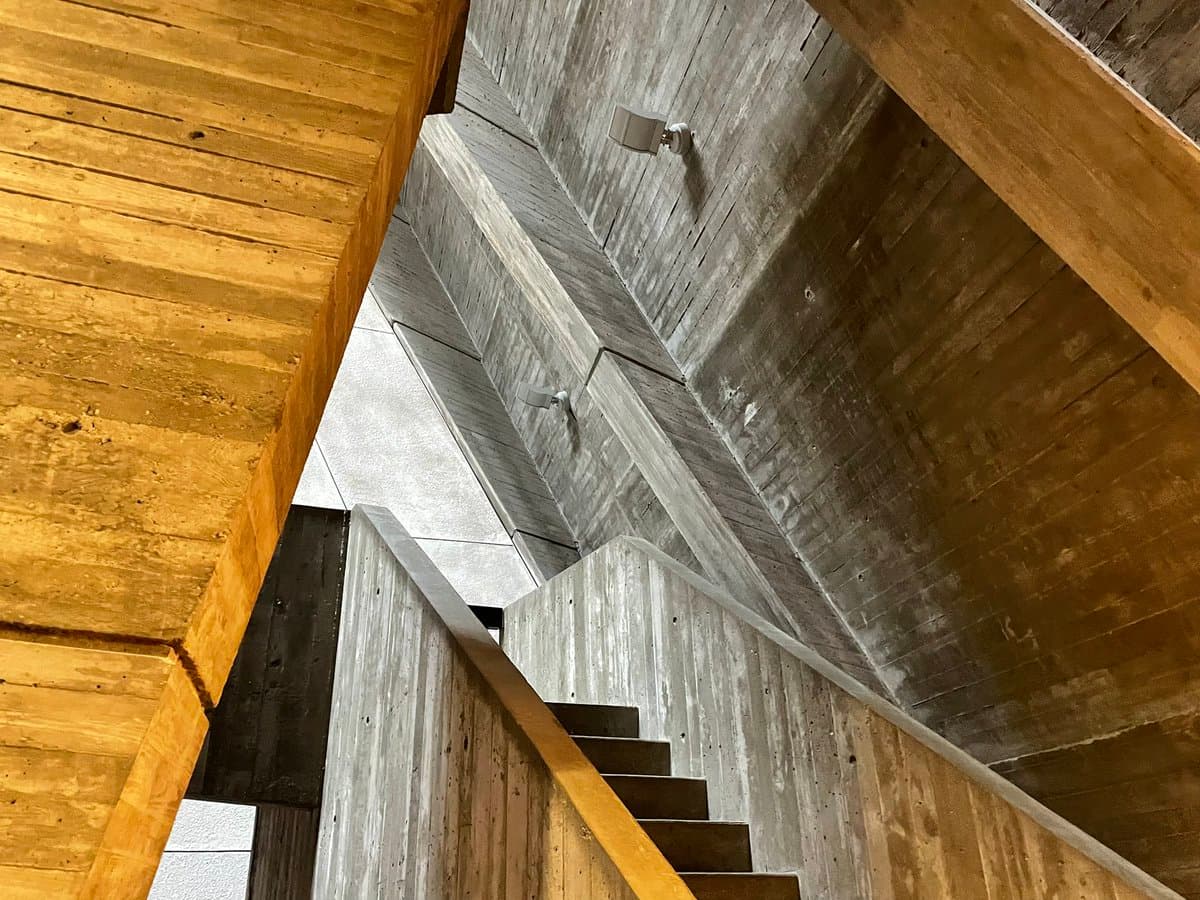 A low-angle architectural photograph looking up a concrete staircase with a wooden handrail, showing the textured concrete walls and ceiling of a brutalist structure.