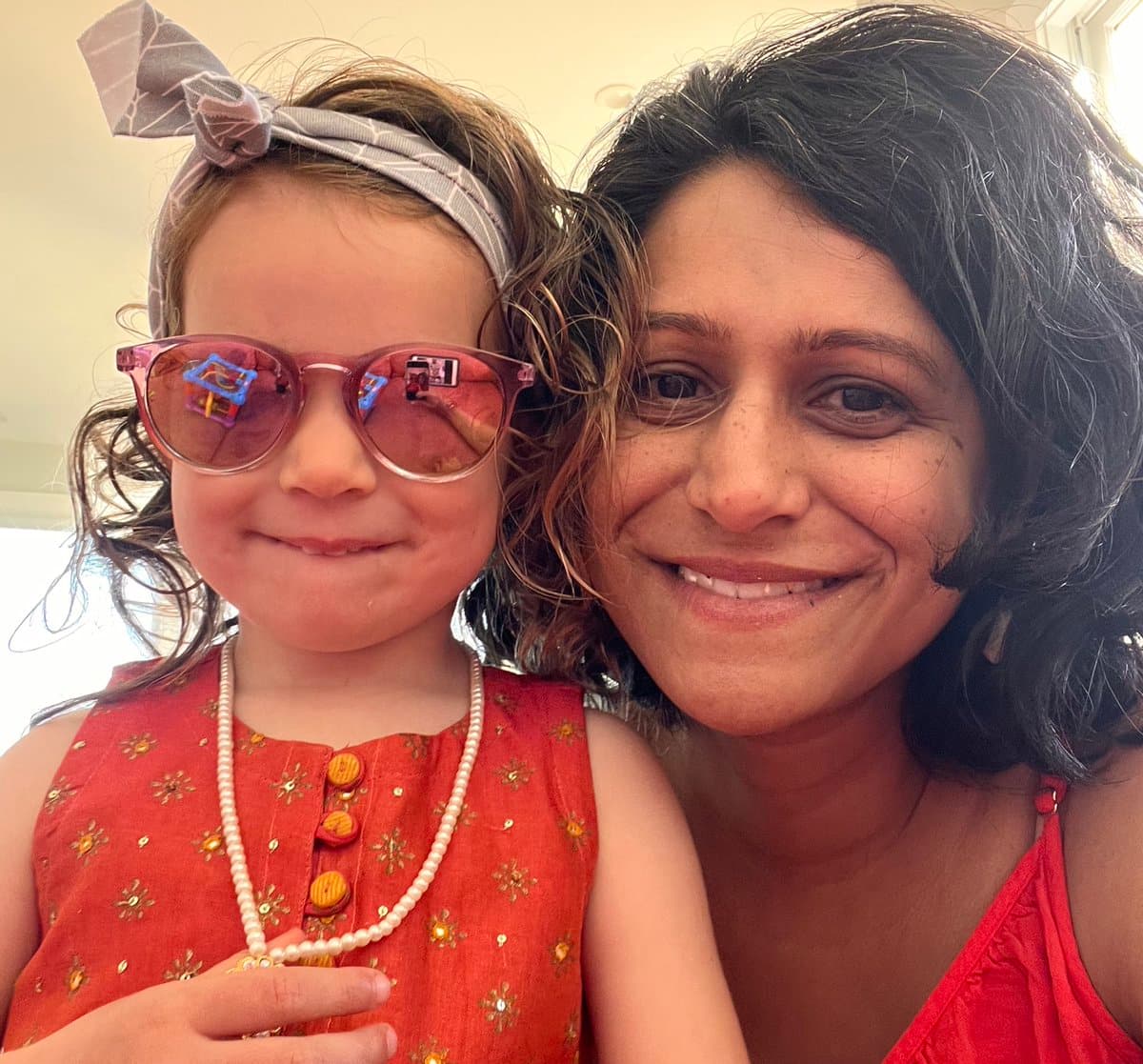 A selfie of a woman and a young girl posing together, both with traces of colored powder on their faces and hair, consistent with a Holi celebration. The girl wears pink sunglasses, a headband, and a red patterned top, while the woman wears a red top.