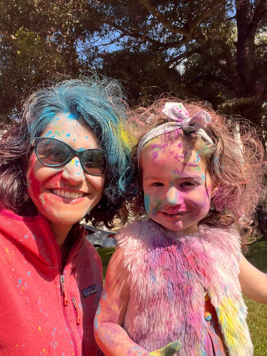 A woman and a young girl pose for a selfie outdoors, both covered in colorful Holi powder on their faces, hair, and clothing. The woman wears sunglasses and a red fleece jacket, while the girl wears a fluffy pink vest and a headband with a bow. They are smiling in front of a backdrop of trees and a parked car.
