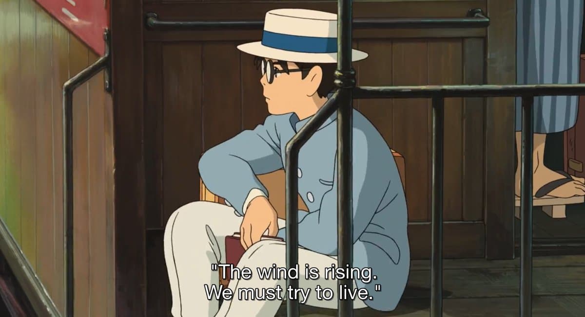 A screenshot from an animated film showing a young man in a light blue suit, white hat, and glasses sitting on a train platform. He is looking to the left with a serious expression.