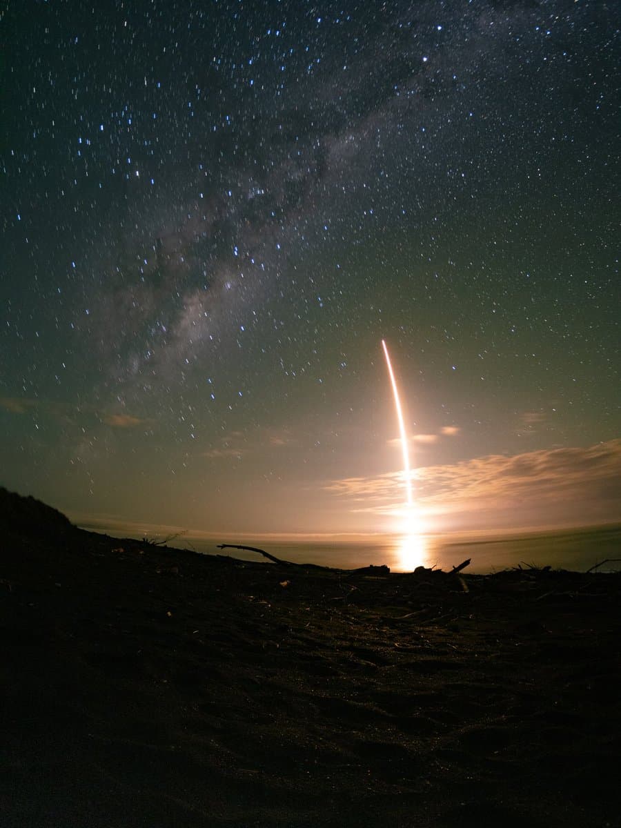 A long-exposure night photograph showing a bright, vertical light trail from a rocket launch rising over the ocean horizon, set against a starry sky with the Milky Way visible.