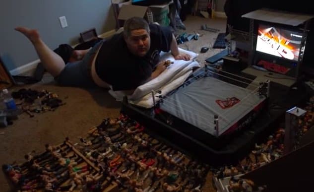 A man lies on the floor next to a miniature wrestling ring surrounded by numerous action figures, with a computer monitor displaying a wrestling game in the background.