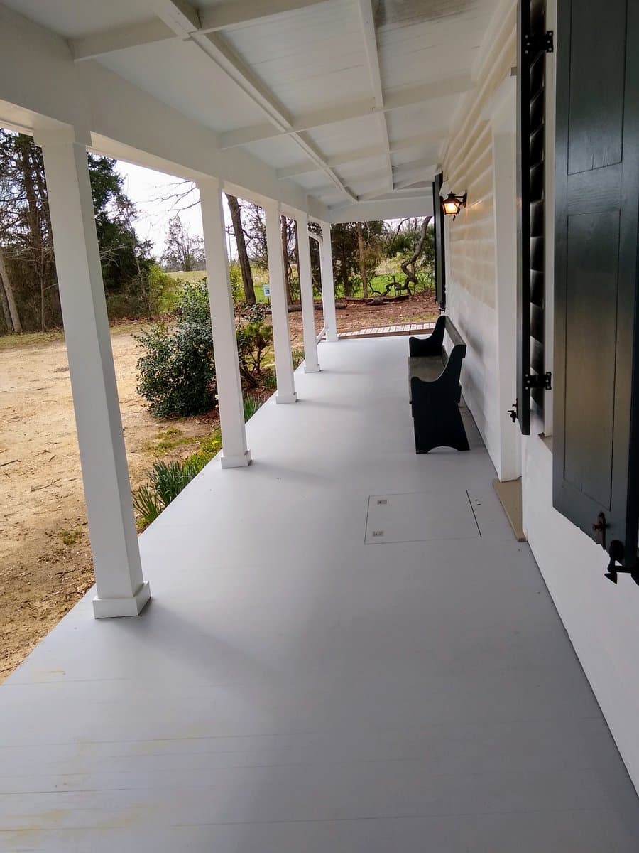 A long, covered porch with white pillars and a light gray floor, featuring a black bench and a wall-mounted light fixture. The porch overlooks a natural landscape with trees and shrubs.