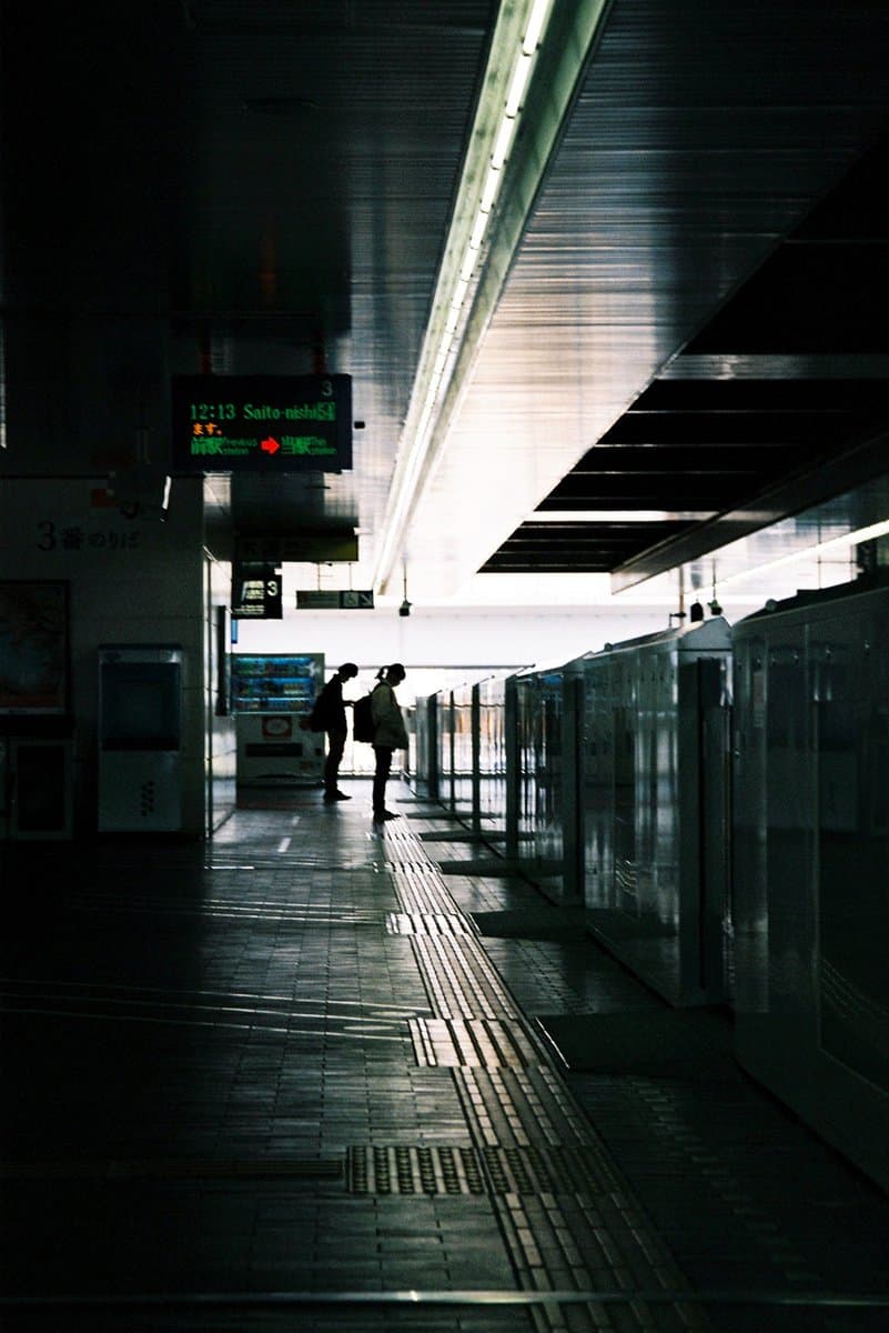 A moody, high-contrast photograph of a train station platform with two silhouetted figures standing near the edge. A digital departure board hangs overhead, and the scene is illuminated by a bright strip of light from the ceiling.