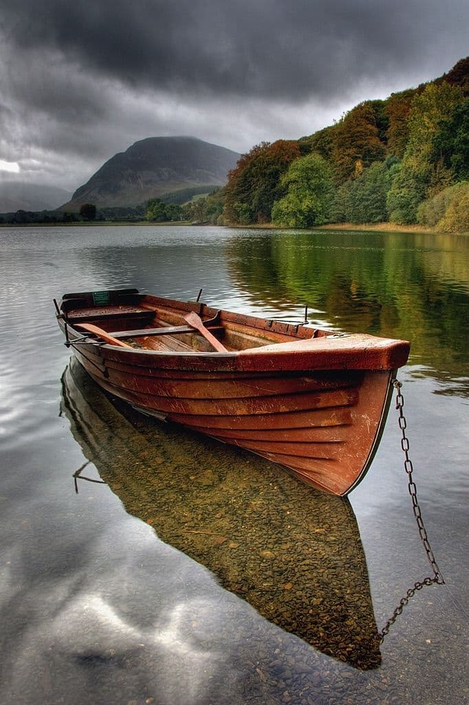A weathered wooden rowboat floats on a calm lake, moored by a chain, with a forested hill and a mountain under a cloudy sky in the background. The boat's reflection is clearly visible on the water's surface. There is no legible text in the image.