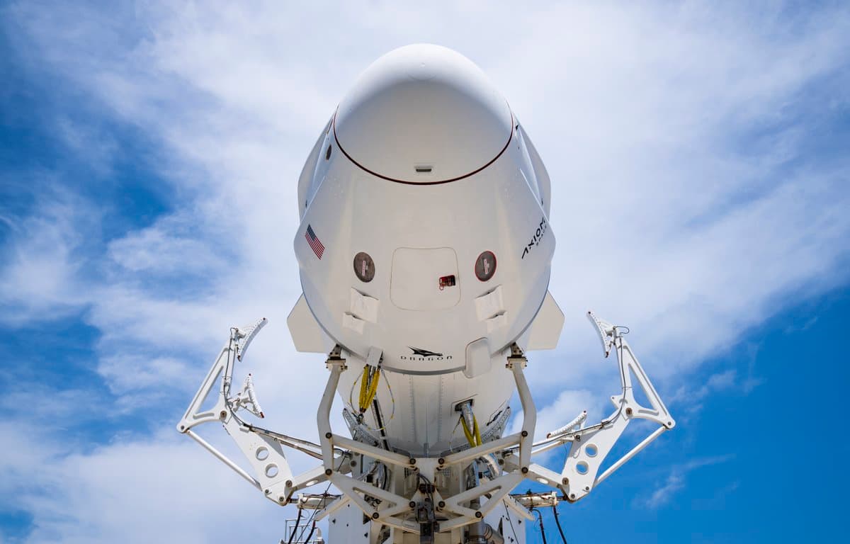 A low-angle photograph of a white SpaceX Falcon 9 rocket and Dragon capsule mounted on a launch tower against a blue sky with scattered clouds. The capsule features the Axiom Space logo and an American flag, with the word "DRAGON" visible on its lower section.