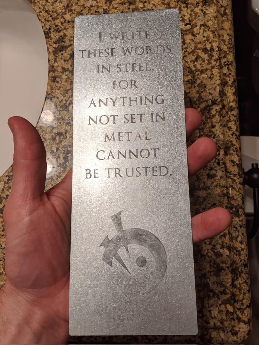 A person holds a rectangular metal ingot engraved with a quote and a logo, set against a granite countertop.