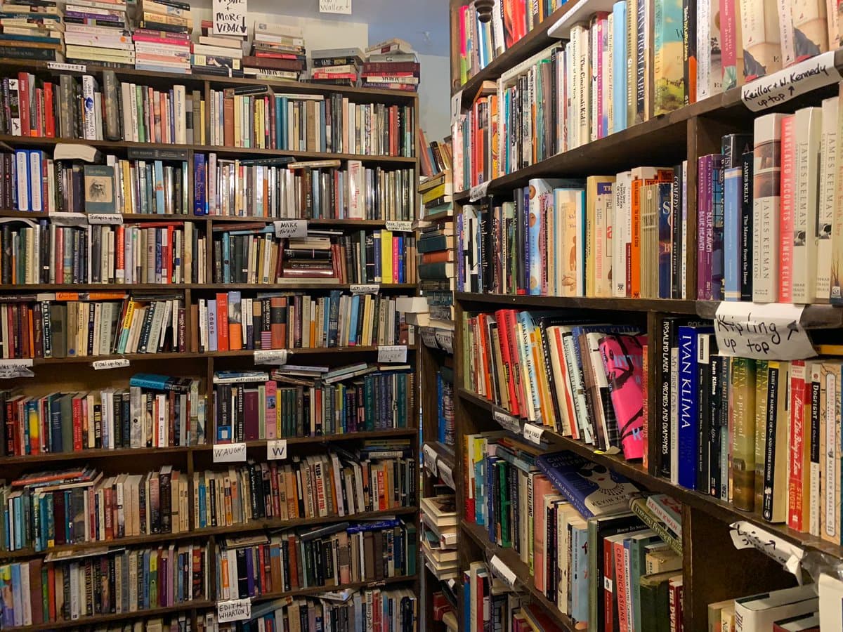 A photograph of a crowded room filled with floor-to-ceiling wooden bookshelves packed with numerous books. Handwritten paper labels are attached to the shelves to organize the collection by author or title.
