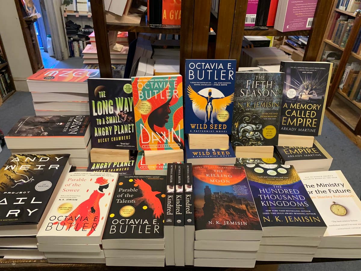 A display of numerous science fiction and fantasy books arranged on a table in a bookstore, featuring prominent titles by authors such as Octavia E. Butler, N.K. Jemisin, and Andy Weir. The books are stacked in piles and propped up on wooden stands to showcase their covers.
