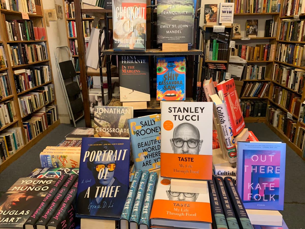 A display of new and used books arranged on tables and shelves inside a bookstore, featuring titles such as 'Taste' by Stanley Tucci, 'Portrait of a Thief' by Grace D. Li, and 'Out There' by Kate Folk.