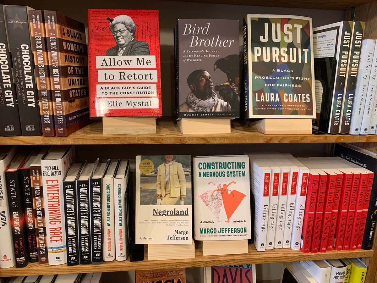 A photograph of a bookshelf displaying several books, with three titles prominently displayed on wooden stands: 'Allow Me to Retort', 'Bird Brother', and 'Just Pursuit'.