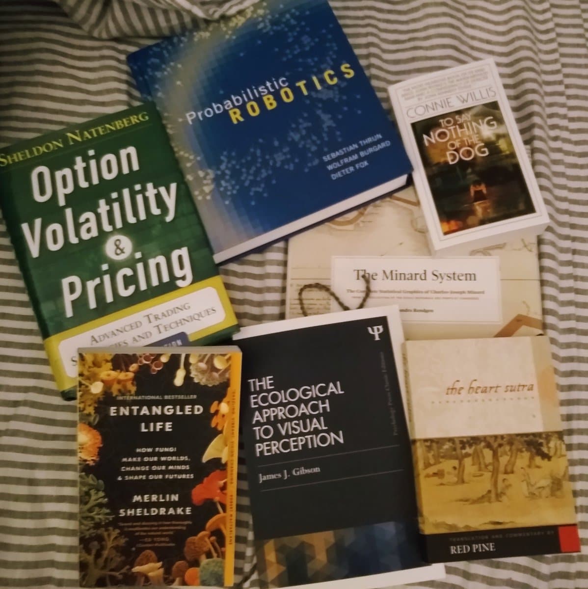 A flat-lay photograph of seven books arranged on a striped bedsheet. The books include titles on finance, robotics, ecology, and fiction.