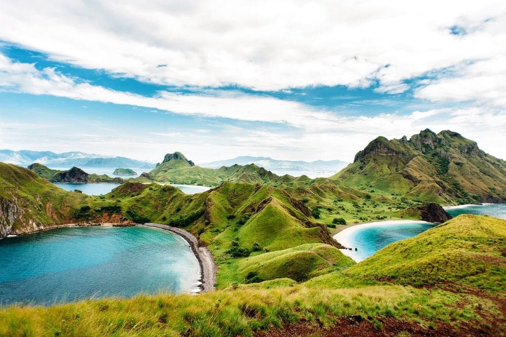 A scenic landscape photograph showing lush green hills and mountains surrounding turquoise bays and beaches under a partly cloudy sky. The view appears to be taken from an elevated vantage point overlooking a coastal archipelago.
