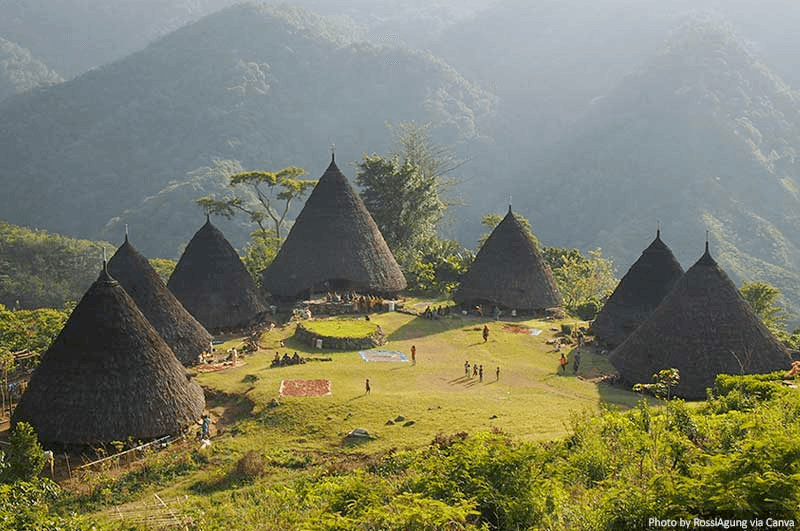 A landscape photograph showing a village of several conical thatched-roof huts on a grassy hillside, surrounded by lush green vegetation and misty mountains in the background. Small figures of people are visible on the grass between the structures.