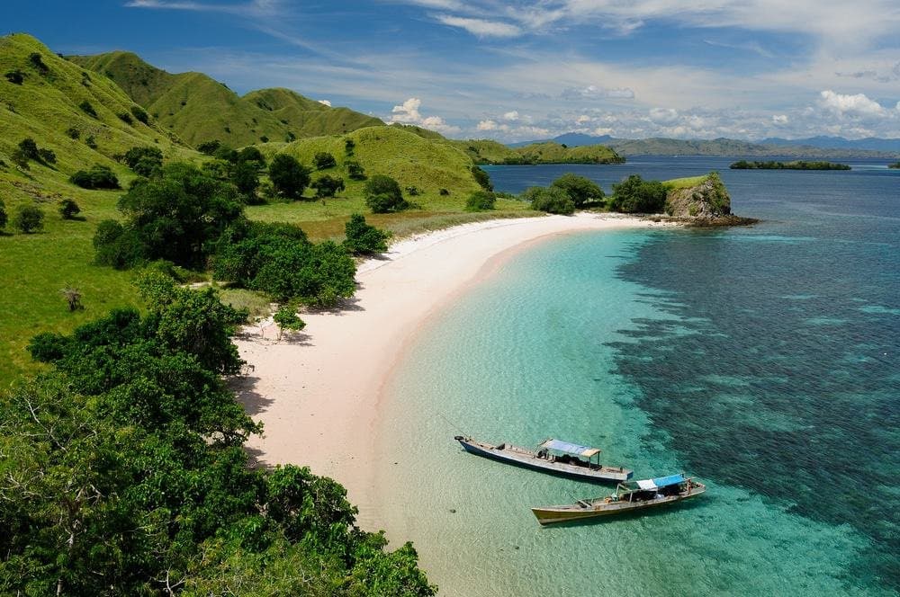 An aerial view of a tropical coastline featuring a white sand beach bordered by lush green hills and turquoise water. Two small boats are anchored in the shallow water near the shore.