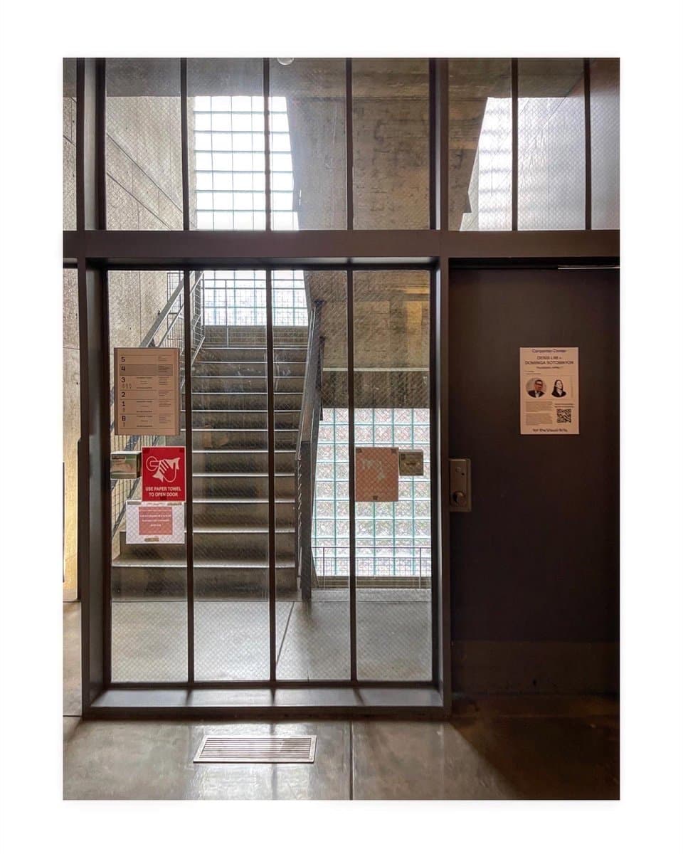 A photograph of a modern building interior featuring a glass and metal entrance gate that looks out onto a concrete staircase. The scene includes a dark brown door on the right and several informational signs attached to the glass panels.