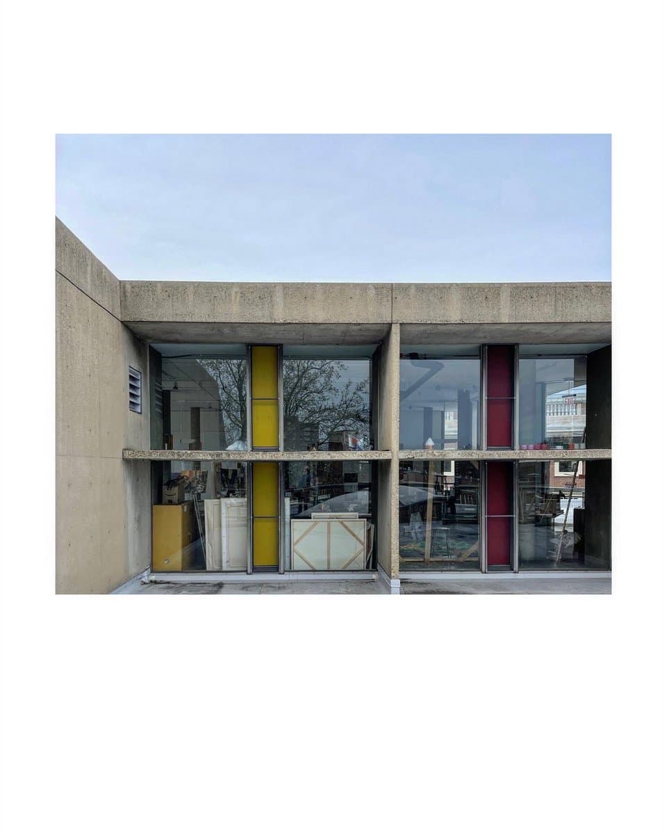 A photograph of the exterior of a modern building, featuring a concrete facade with large glass windows. The windows reveal an interior with yellow and red vertical panels, along with various items and furniture, suggesting a workspace or gallery space.